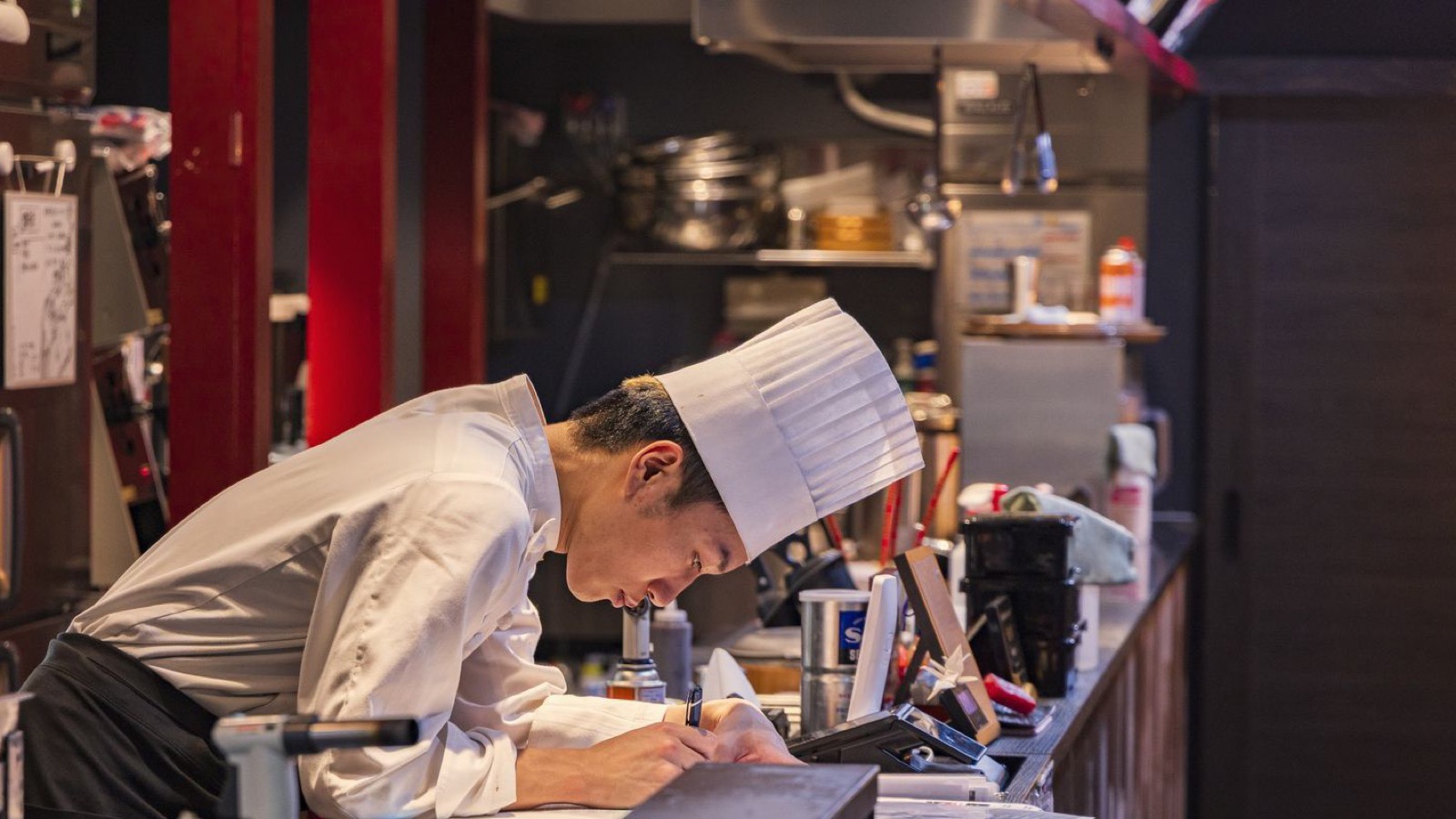 A cook writing notes at the pass at the end of service, the habit a shift report template turns into a record